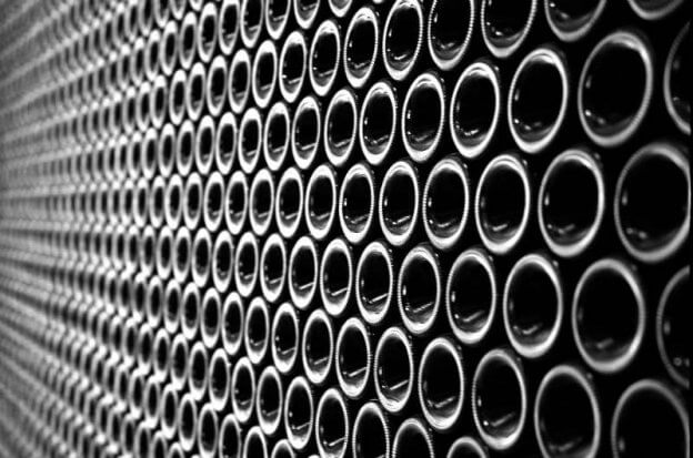 Fotografia in bianco e nero di un "muro di bottiglie". Le bottiglie sono disposte distese du un fianco in molte file sovrapposte. La foto inquadra il fondo delle bottiglie